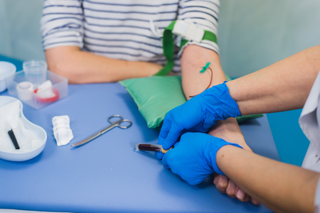 Preparation for blood test with pretty young blond woman by female doctor in white coat medical uniform on the table in white bright room. Nurse pierces the patients arm vein with needle blank tube.の写真素材