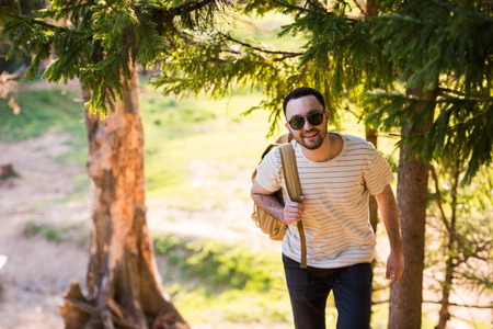 Handsome stylish young man wearing white t-shirt and sunglasses with backpack in his hand is...