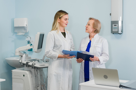 Senior woman doctor and beautiful young female doctor in white medical coats are talking and smiling...