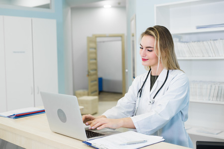 Smiling female doctor wearing a scrub and working at the hospital reception, she is writing a medical report on a clipboard and scheduling appointmentsの写真素材