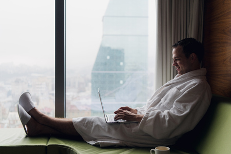 Young smiling businessman working on laptop computer wearing white bath robe sitting near window with cup of coffee looking at sunrise city view. Motivation conceptの写真素材