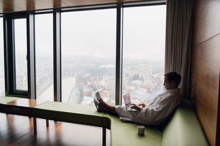Young smiling businessman working on laptop computer wearing white bath robe sitting near window with cup of coffee looking at sunrise city view. Motivation conceptの写真素材