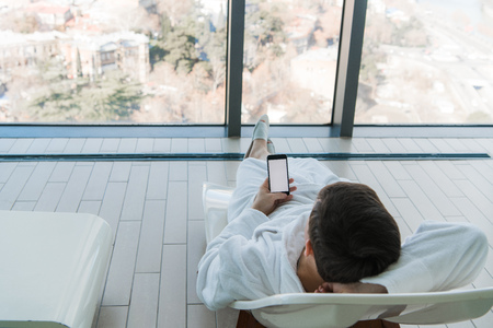 Young man lying on sunbed near pool with phone indoors. Luxury hotel with a great city view behind...