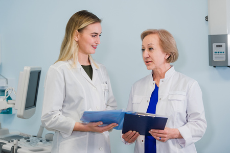 Senior doctor talking with young woman assistant standing in the gynecological office with chair and lamp on the backgroundの写真素材