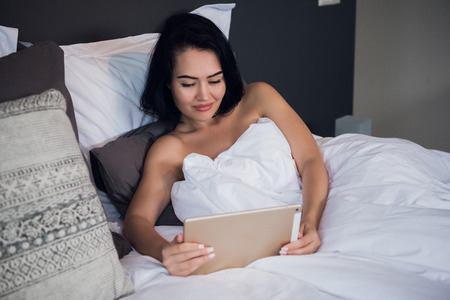 Happy young woman greeting in a tablet video call on line sitting on bed in the bedroom at homeの写真素材