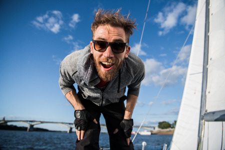 Attractive man in sunglasses on the yacht. Close portrait of a happy crazy face, attractive sportsman yachstman laughing. Close up outdoor portrait of sailor, strong man on a boat on river or sea.の写真素材