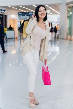 Beautiful girl in shopping mall, looking at camera, smiling widely and holding colorful shopping bags in raised hands. Turned a little bit aside. Wearing white jeansの写真素材