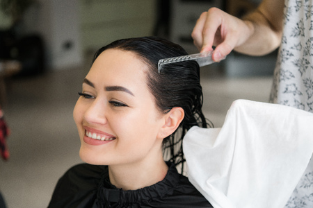 Close up of a young happy beautiful woman smiling to the camera while professional hairdresser wrapping her wet hair in a towel after washing it with shampooの写真素材