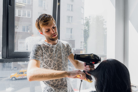 Professional smiling male stylist blow drying womans hair with a dryer in salon