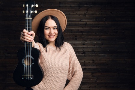 Young funny hipster girl having fun and playing on small ukulele guitar, singing and dancing. wearing vintage hat, joy, positive mood. Dark wooden wall background.の写真素材
