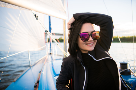 Young smiling dark-haired woman wearing sunglasses sitting on a board of boat in sunny autumn day, walking on a sea or riverの写真素材
