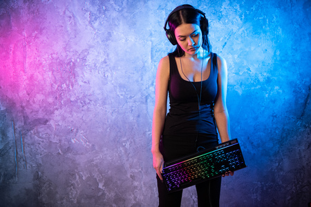 Young Sexy Disco Girl Wearing Gaming Headset Holding Gaming Kyeboard. Room Lit in Retro, Retrowave...
