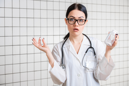 Surprised female doctor gesturing with hands and pills while standing with mouth open on medical office background.の写真素材