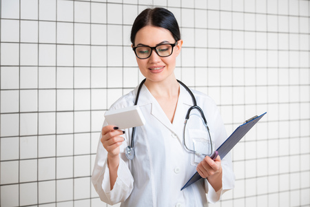 A young thin dark-haired girl with glasses, dressed in a medical overall, reads something on the pills pack next to the office background in the pharmacy.の写真素材