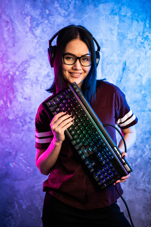 Nerd geek young adult women holding gaming keyboard over colorful pink and blue neon lit wall. Gaming gamers concept.の写真素材