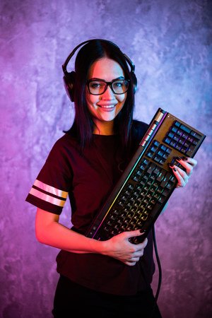 Beautiful Friendly Pro Gamer Streamer Girl Posing With a Keyboard in Her Hands, Wearing Glasses. Attractive Geek Girl with Cool Neon Retro Colors in Background.の写真素材