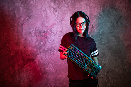 Portrait of the Beautiful Young Pro Gamer Girl standing with a gaming keyboard and headset and Looks...