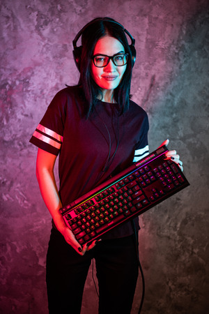 Beautiful Friendly Pro Gamer Streamer Girl Posing With a Keyboard in Her Hands, Wearing Glasses. Attractive Geek Girl with Cool Neon Retro Colors in Background.の写真素材