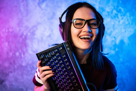 Beautiful Friendly Pro Gamer Streamer Girl Posing With a Keyboard in Her Hands, Wearing Glasses. Attractive Geek Girl with Cool Neon Retro Colors in Background.の写真素材