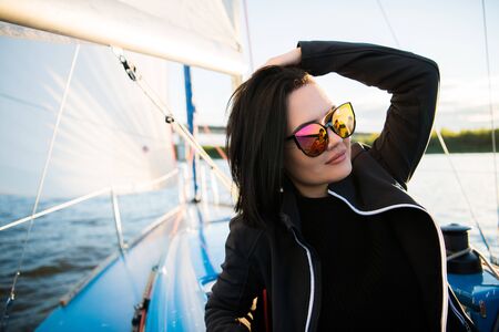 Beautiful young woman sits on yacht bowl and poses. She wears sunglusses with hand and smile. Model is sailing on board of yacht.の写真素材