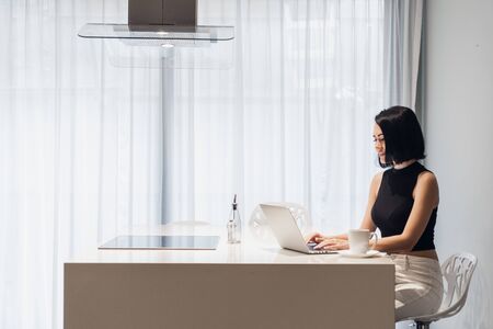 Beautiful business woman is using a laptop, looking at camera and smiling while sitting at homeの写真素材