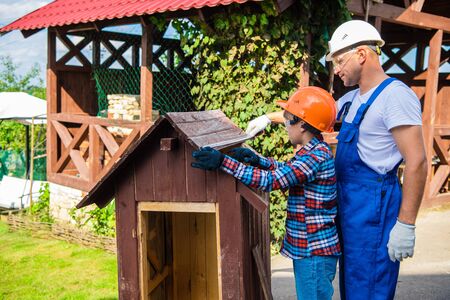 Boy, teenager busy in protective helmet prepairing to repair the dog house with his dad. Father teaching little son to properly measure with a tape-line.の写真素材