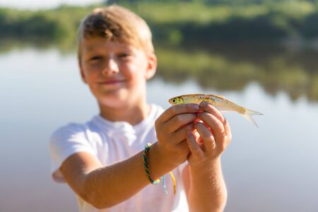 A teen boy enjoying catch. Young blond boy holding a fish outside in sunny summer day. Childhood and...