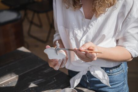Female potter sitting and stirs paint with a brush a cup on the table. Woman making ceramic item. Pottery working, handmade and creative skills.の写真素材