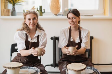 Two women make pottery on a pottery wheels, shaping clay by their hands, close up. Pottery craft workshop at art studio.の写真素材