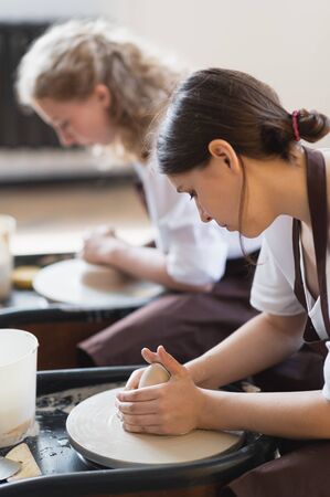 Two women make pottery on a pottery wheels, shaping clay by their hands, close up. Pottery craft workshop at art studio.の写真素材
