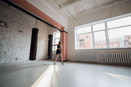 A pretty kickboxer girl is preparing for competitions in the boxing hall, practicing the technique of punches on a punching bagの写真素材