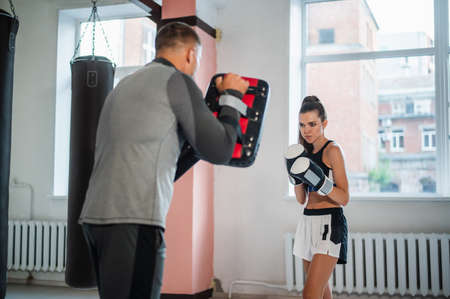 Warm-up before training. A girl in a Boxing stance with gloves is preparing to strike at the paws in the hands of the trainerの写真素材