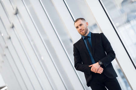 Stylish young businessman wearing a modern suit, who is a high achiever, standing on the top floor of an office building looking out at the view through large windowsの写真素材