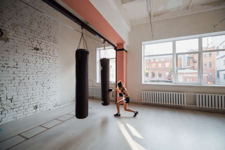 A girl in sports clothes is engaged in boxing and works out a punch with her hand on a punching bagの写真素材