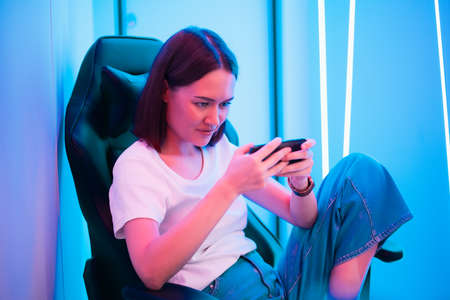 Cute gamer girl sit on a gaming chair, smiling and playing mobile online game on a smartphone. Having fun playing with friends online.の写真素材