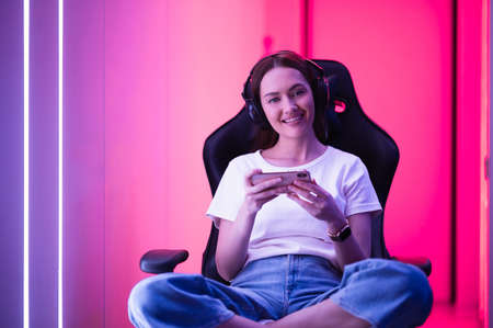 Young girl playing mobile online game on a smart phone while sitting on gaming chair in colorful neon light room.の写真素材