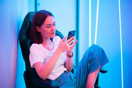 Young lady sitting on gaming chair with cellphone in hands. Playing online mobile game or browsing social networks resting in a room lit with neon color.の写真素材