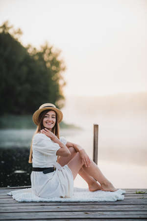Attractive woman wearing white dress and hat sitting near lake. Warm and cozy summer morning atmosphere.の写真素材