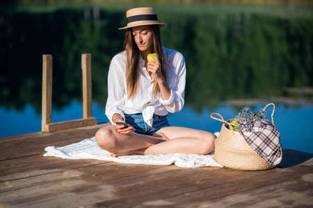Beautiful young woman in hat has picnic near the lake in summer forest. She eats an apple and looking at smart phone.の写真素材