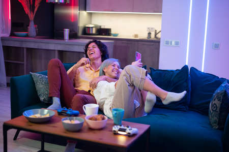 Happy boyfriend and girlfriend laughing after playing video games at home, joysticks and snacks on a...