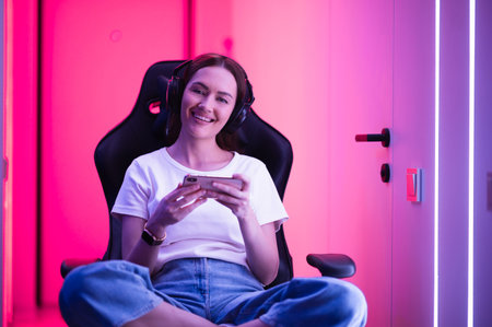 Gamer girl in a big professional music headphones in white t-shirt and blue jeans holds smart phone in hands and plays game app on mobile phone, smiling and looking at camera indoors.の写真素材