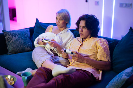 Couple of young adults playing video games at home. Emotional diverse gamers holding joysticks and compete in intense video game on gaming console.の写真素材