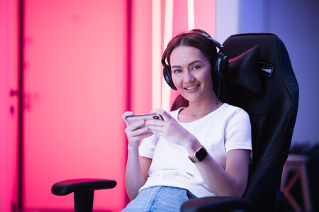 Gamer girl in a big professional music headphones in white t-shirt and blue jeans holds smart phone in hands and plays game app on mobile phone, smiling and looking at camera indoors.の写真素材