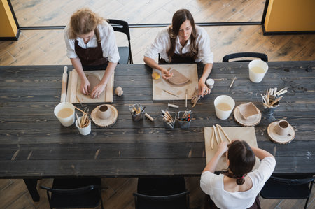 In the clay workshop. Female potters talking and smiling during the working process in the clay workshop. Female potter masters rolling up the clay on table with ceramic products.の写真素材