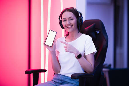 Smiling gamer girl wearing headset showing smart phone and pointing at blank screen with her finger.の写真素材