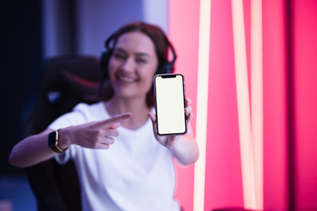 Cheerful gamer girl pointing with finger at smartphone with blank screen while sitting on a gaming chair.の写真素材