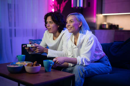 Happy boyfriend and girlfriend playing video games and holding joysticks at home. Millennial couple enjoying game while sitting on sofa and spending free time. Concept of relationshipの写真素材