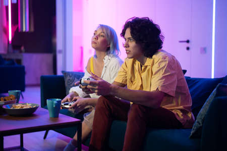 Happy boyfriend and girlfriend playing video games and holding joysticks at home. Millennial couple...