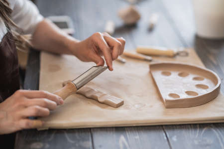 Craftsman hands connect the clay parts made of raw clay, master fingers work with pieces of clay, female sits at a workshop behind the tableの写真素材