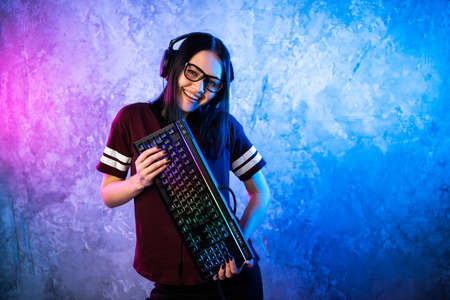 Beautiful Friendly Pro Gamer Streamer Girl Posing With a Keyboard in Her Hands, Wearing Glasses. Attractive Geek Girl with Cool Neon Retro Colors in Background.の写真素材